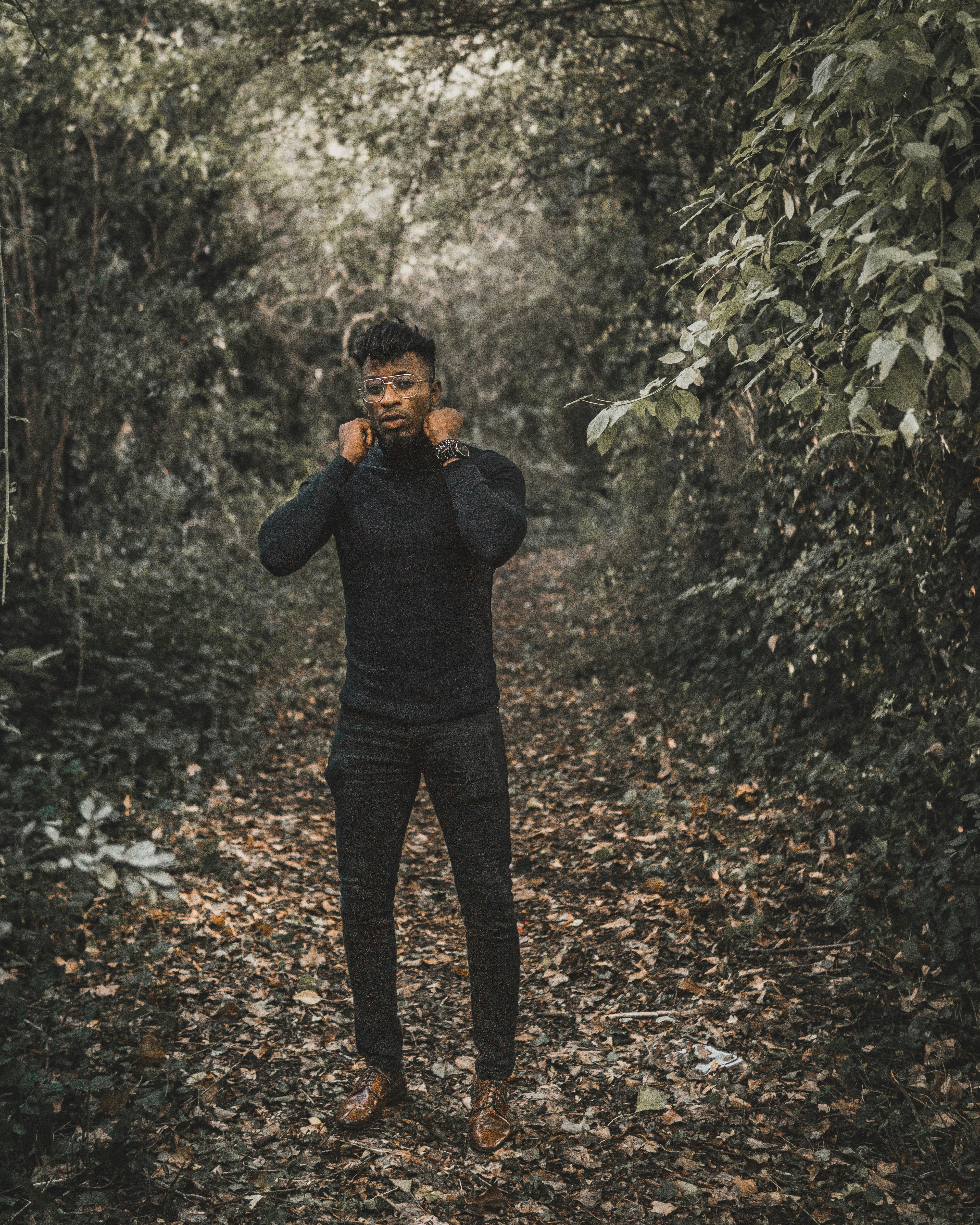 standing man wearing black turtleneck sweater and black fitted pants surrounded with trees during daytime